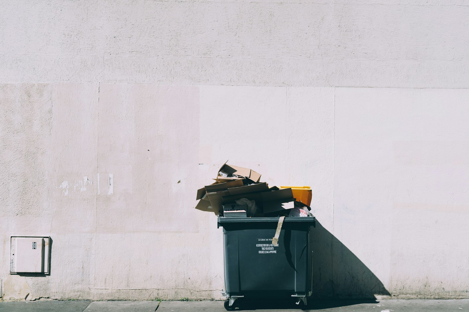 Szary pojemnik na kółkach pod białą ścianą, na wierzchu kartony